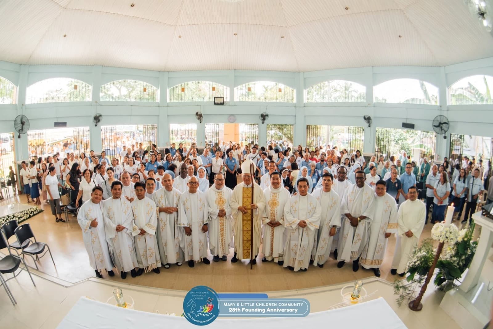 Priests in white vestments and a bishop holding a crozier stand at the front of a bright, crowded chapel, with sisters and members of the Community filling the pews behind them, at the 28th founding anniversary Mass