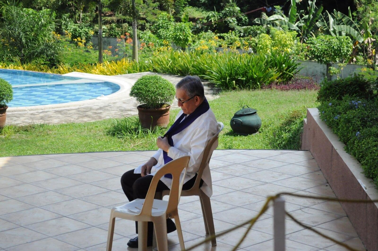 A priest wearing a purple stole, sitting on a chair in a garden beside a swimming pool