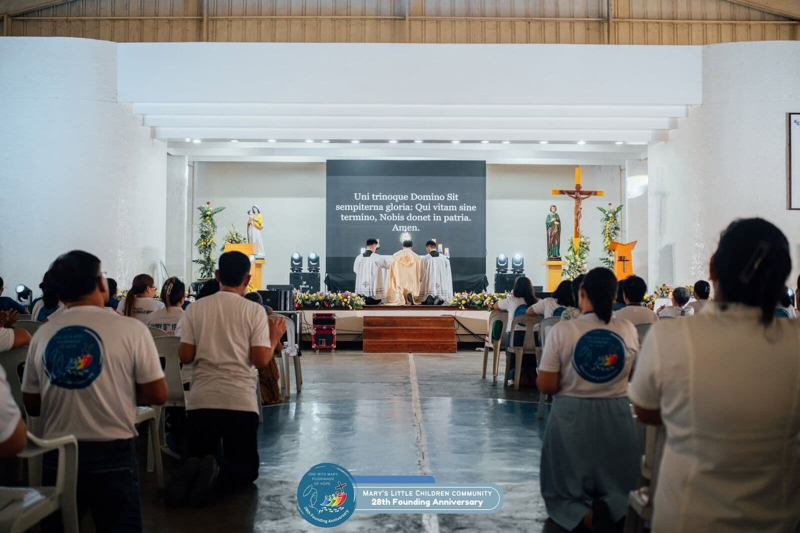 Members in Community shirts kneeling at Mass in a large hall, the priests at the altar beneath a screen of Latin prayer