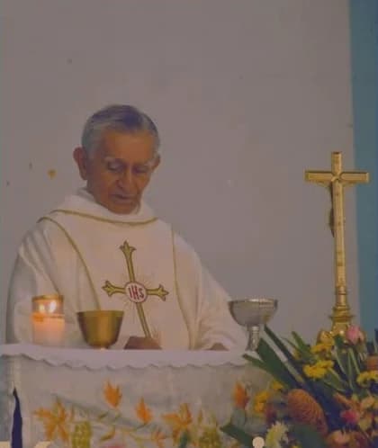 Msgr. Fred, white-haired, in white vestments, celebrating Mass at an altar with a chalice, a candle and a brass crucifix, flowers in front.